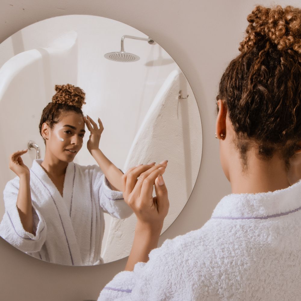 Woman in a white robe applying cream to her face in front of a round mirror.