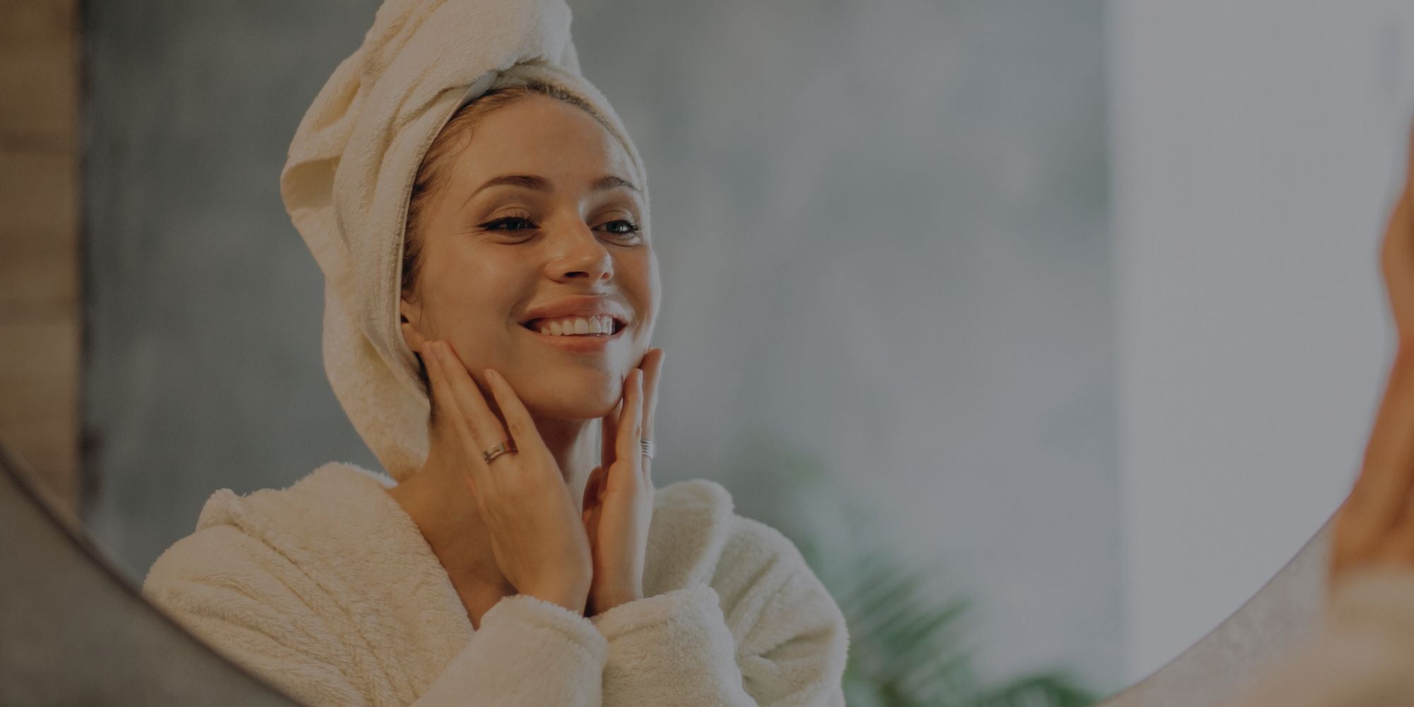 Woman in a white robe with a towel wrapped around her head, smiling and touching her face.