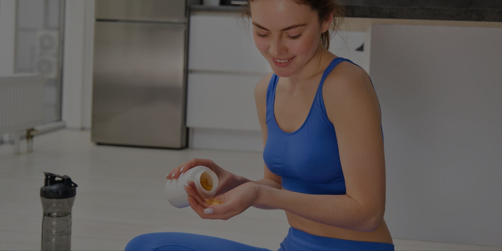 Woman in blue athletic wear sitting on the floor with a supplement bottle in a kitchen.
